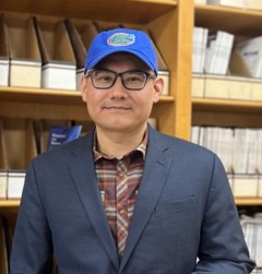 photo of Xiaofei Bai smiling directly at the camera. He is wearing black glasses, a gray blazer over a plaid shirt, and a blue cap with the Florida Gators logo on it. He is standing in front of a bookshelf that contains books and folders.